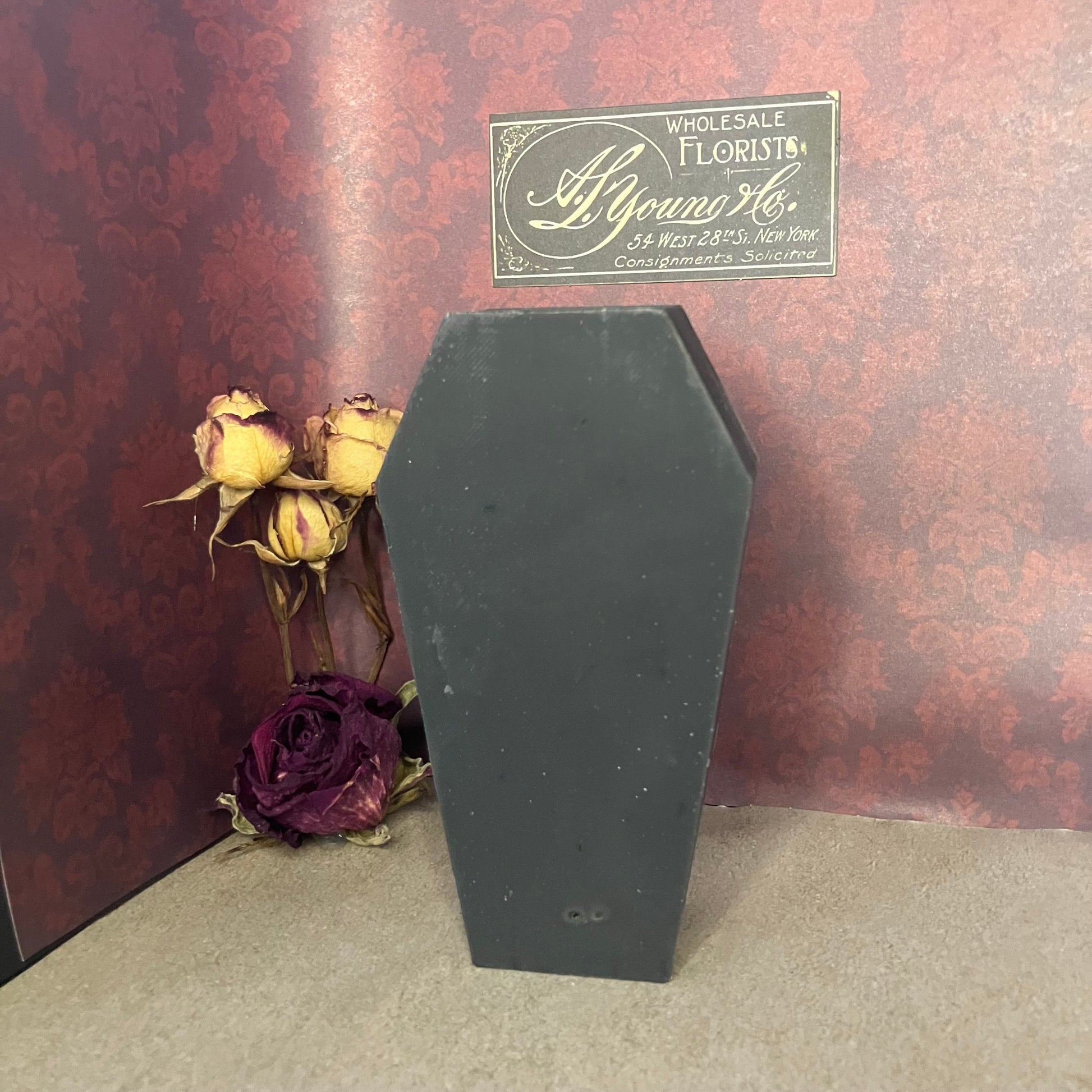 Black coffin-shaped soap with dried roses against a decorative wall.