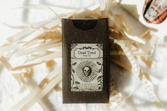 A small black box labeled 'Dead Tired' with a skull illustration, placed on white paper shred with a small round decorative seal nearby.